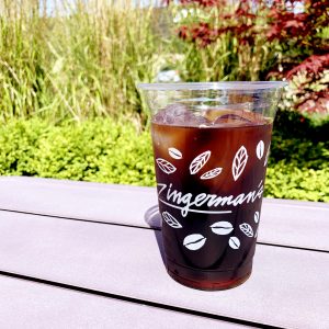 Plastic logo cup filled with ice and dark Zingerman's cold brew coffee, placed upon lavender picnic table with a foliage background